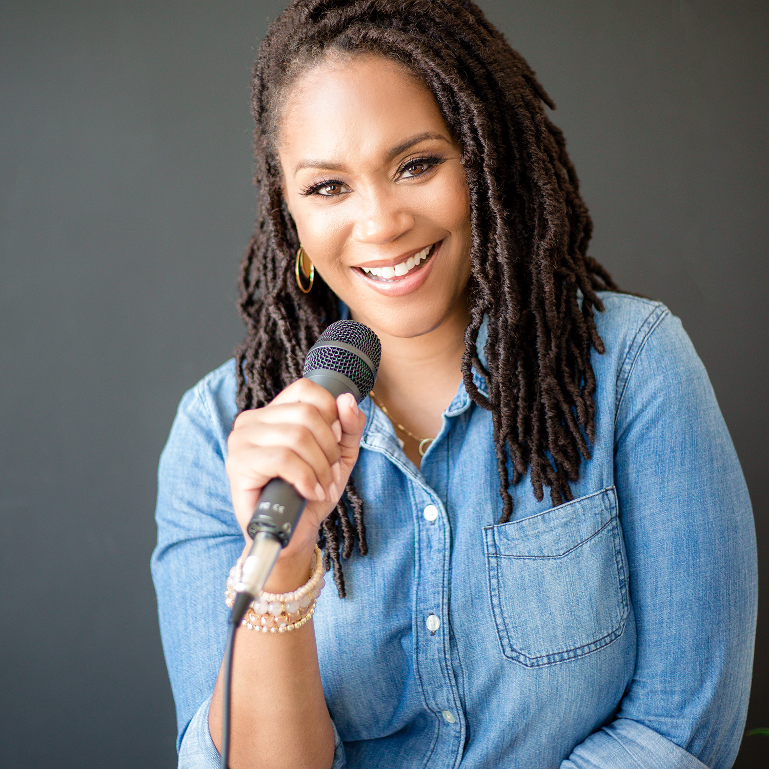Karie Williams headshot in denim shirt against gray background holding a microphone
